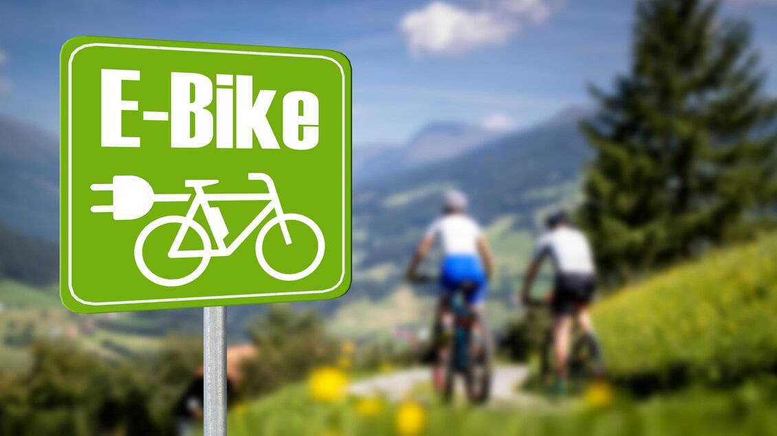 Ein Schild für E-Bikes steht in einer malerischen Landschaft. Im Hintergrund fahren zwei Radfahrer auf einem Fahrradweg. | © TVB Murau