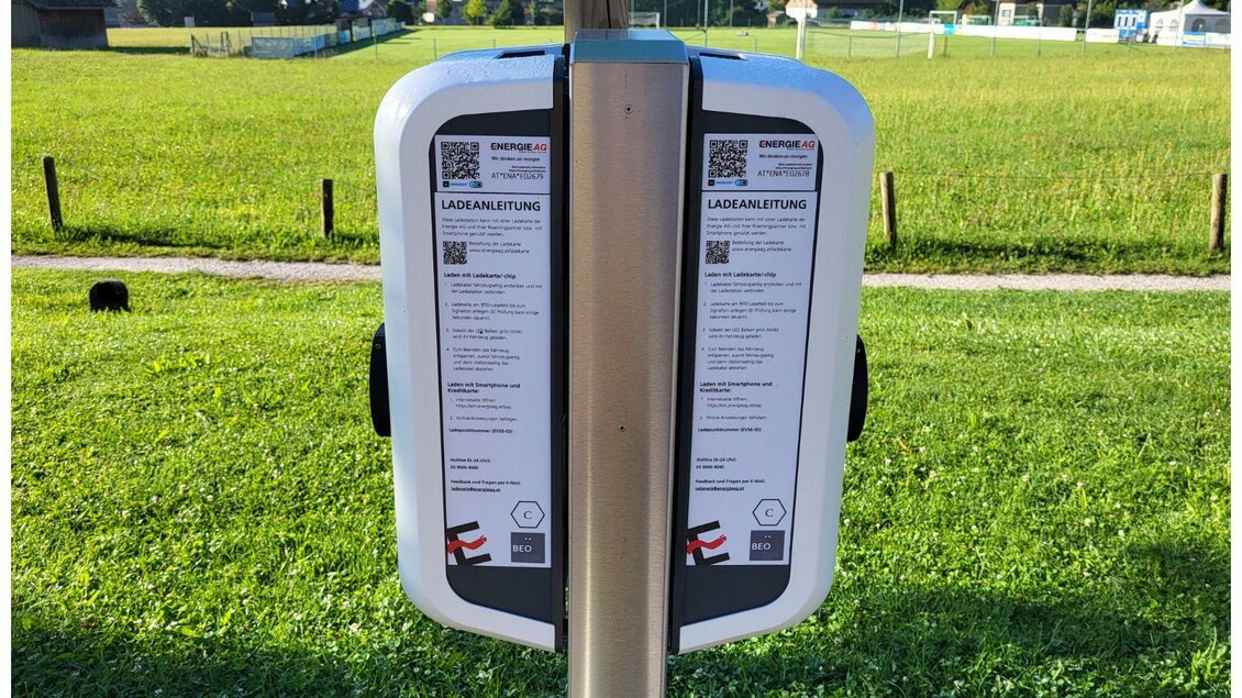 Eine Ladestation für Elektroautos steht auf einem grünen Feld. Im Hintergrund sind Häuser und ein Sportplatz sichtbar. | © Petra Kirchschlager