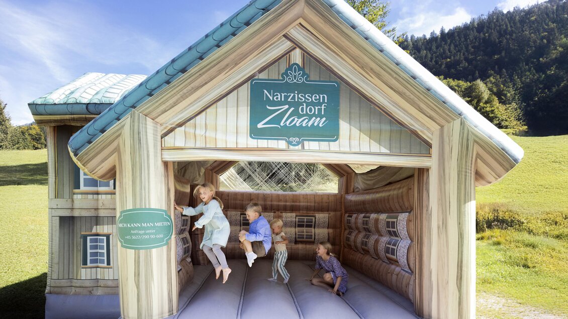 Ein aufblasbares Spielhaus mit dem Schriftzug "Narzissen Dorf Zloam". Kinder spielen und hüpfen darin, umgeben von grüner Wiese und Bergen. | © www.zloam.at