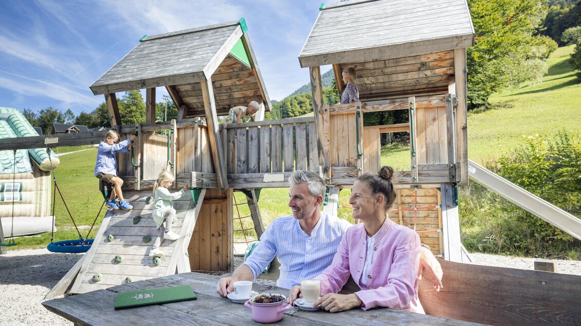Zwei Erwachsene sitzen an einem Tisch in einem Spielplatzbereich. Im Hintergrund spielen Kinder in hölzernen Spielhäusern und auf einer Rutsche. | © www.zloam.at