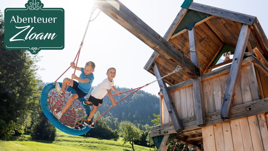 Zwei Kinder spielen fröhlich auf einer großen Schaukel neben einem Holzturm. Die Umgebung ist grün und sonnig, ideal für Abenteuer und Spaß im Freien. | © www.zloam.at