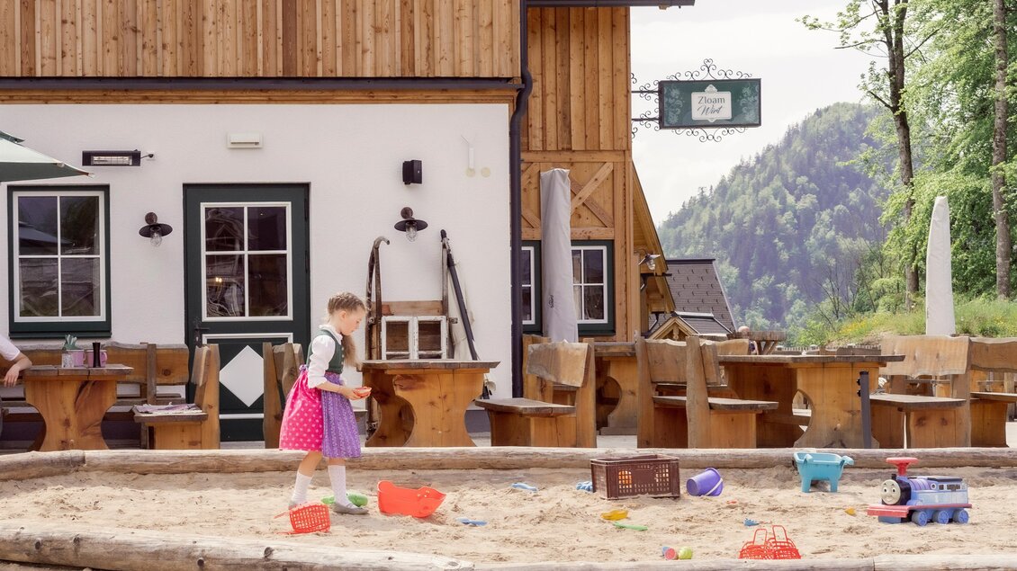 Ein Kind spielt im Sandkasten vor einem Restaurant. Im Hintergrund sind Holztische und Stühle sowie Bäume zu sehen. | © Narzissendorf Zloam, www.zloam.at