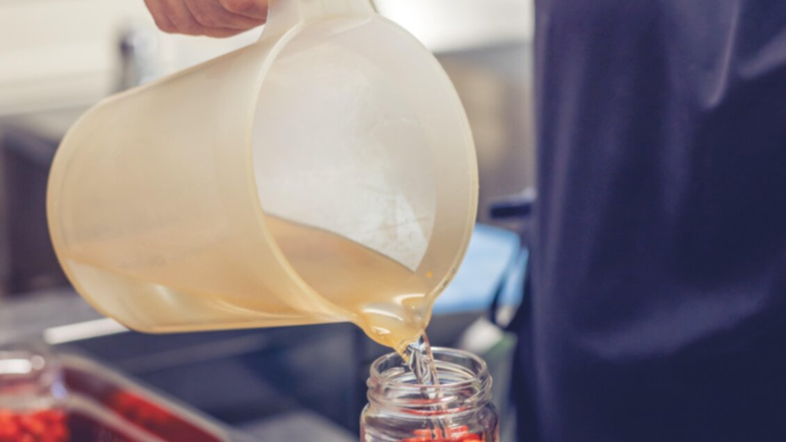 Eine Person gießt eine Flüssigkeit in ein Glas mit roten Kugeln. Die Gläser stehen auf einer digitalen Waage in einer Küchenumgebung.