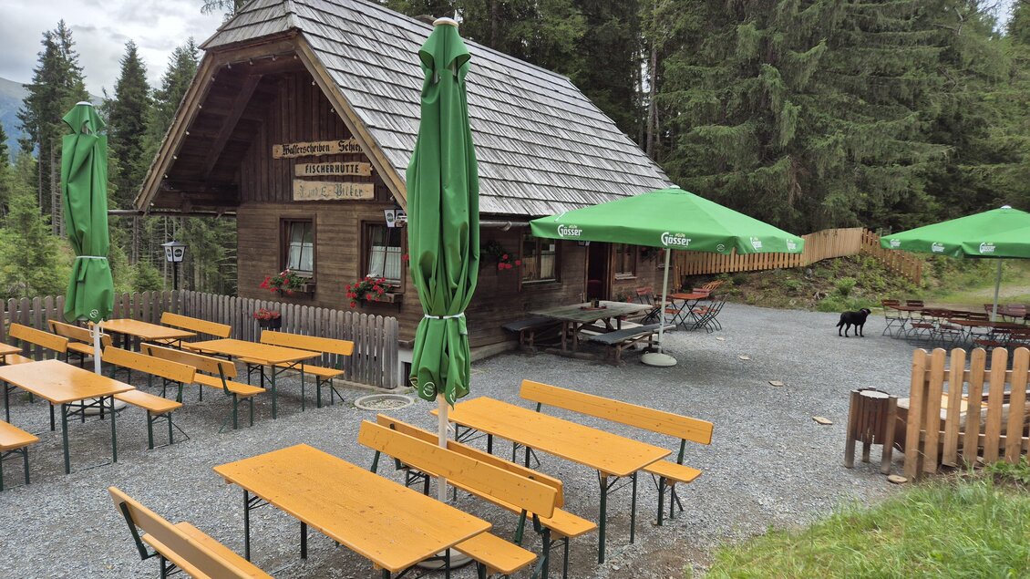 Eine gemütliche Berghütte umgeben von hohen Bäumen. Auf der Terrasse stehen mehrere Holztische mit grünen Sonnenschirmen. | © TVB Murau
