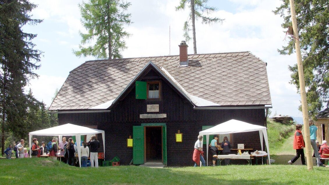 Ein traditionelles schwarzes Holzhaus mit grünen Fensterläden. Vor dem Haus stehen Zelte und Menschen, die an einem Event teilnehmen. | © Naturfreunde