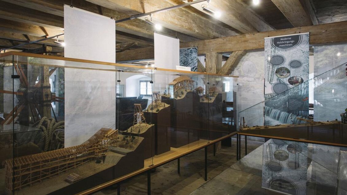 Ein Ausstellungsraum mit mehreren Glasvitrinen, die Modelle und Gegenstände zeigen. Holzdecken und -böden schaffen eine rustikale Atmosphäre. | © Stefan Leitner