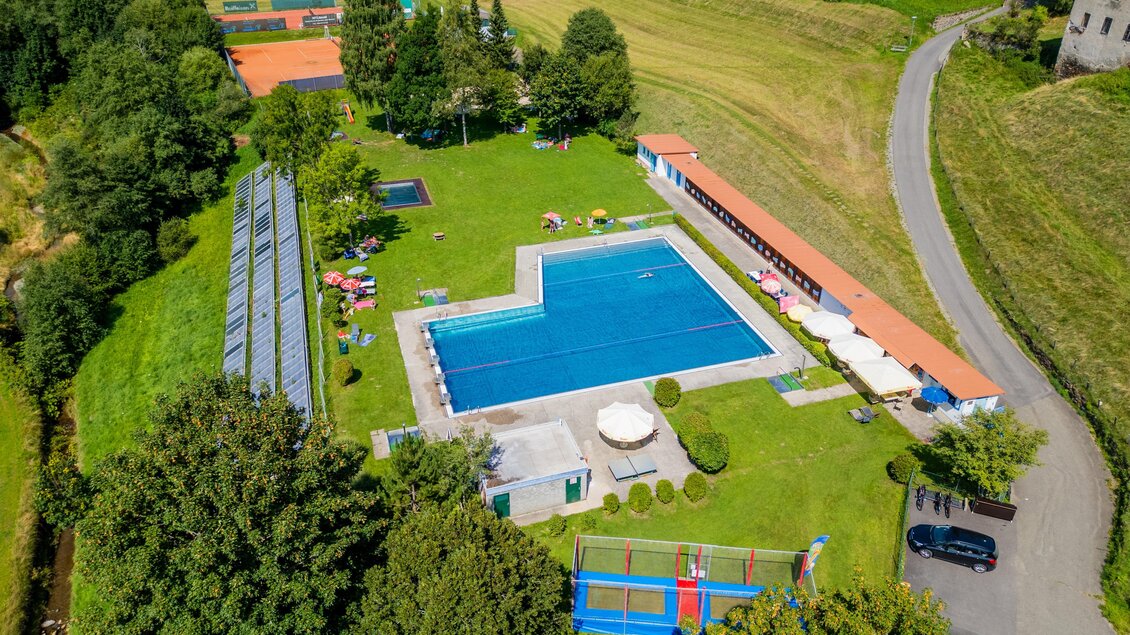 Ein großes Schwimmbad umgeben von grünem Rasen und Bäumen. Im Hintergrund sind Tennisplätze und ein ruhiger Weg zu sehen. | © TV Murau