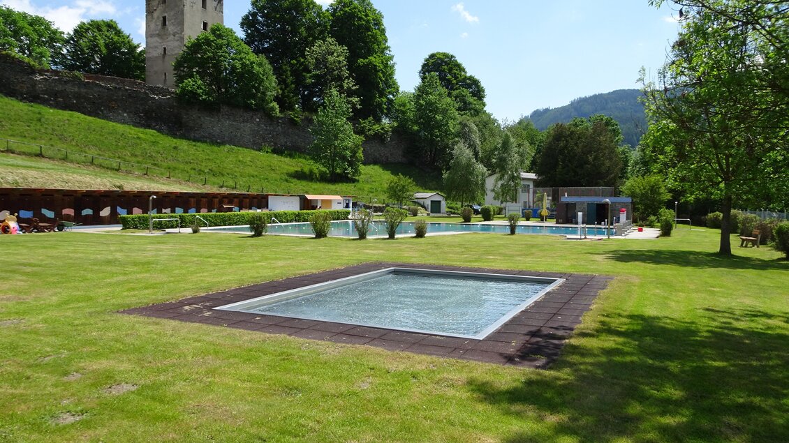 Ein ruhiger Garten mit einem Pool, umgeben von Rasen und Bäumen. Im Hintergrund ist ein alter Turm und ein weiteres Gebäude sichtbar. | © Gemeinde St. Lambrecht