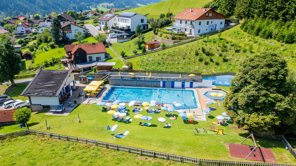 Ein Schwimmbad umgeben von Wiesen und Bäumen, mit Sonnenschirmen und Liegen. Im Hintergrund sind Häuser und die hügelige Landschaft zu sehen. | © TV Murau
