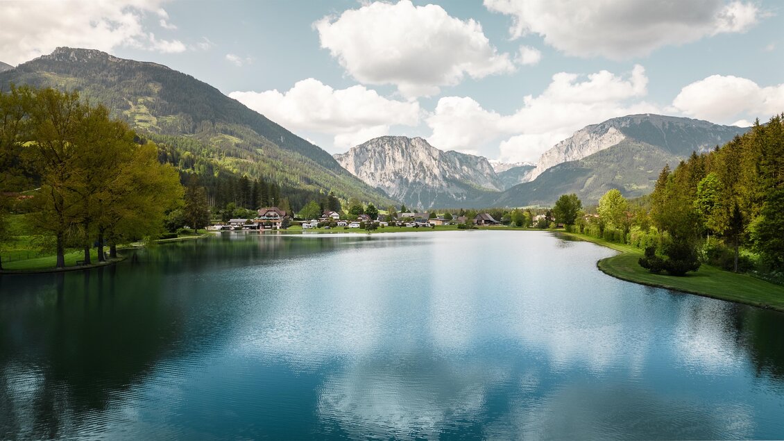 Eine friedliche Landschaft mit einem klaren See und majestätischen Bergen im Hintergrund. Saftige Bäume umgeben das Wasser und der Himmel ist teils bewölkt. | © Hannes Zenz
