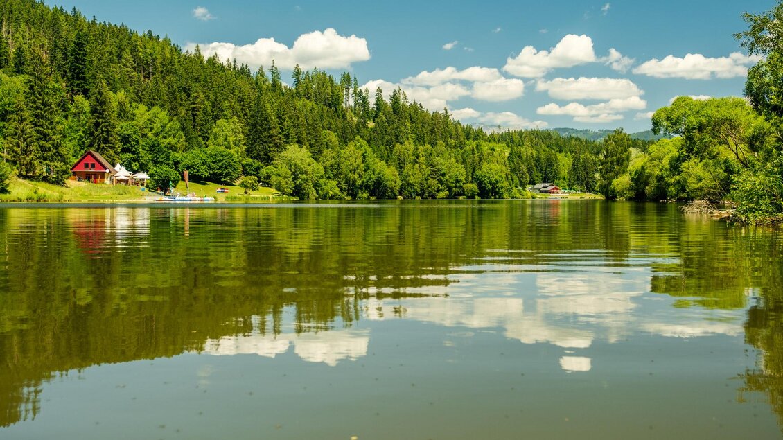 Ein ruhiger See umgeben von grünen Bäumen und Hügeln. Im Hintergrund ist ein rotes Holzhaus zu sehen, das sich im Wasser spiegelt. | © TVB Erzberg-Leoben