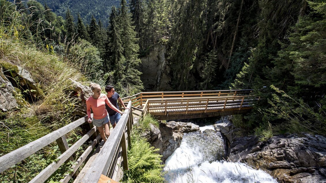 Ein schöner Wanderweg führt zu einem Wasserfall. Zwei Personen genießen die Natur zwischen Bäumen und einem klaren Wasserfluss. | © Holzwelt Murau