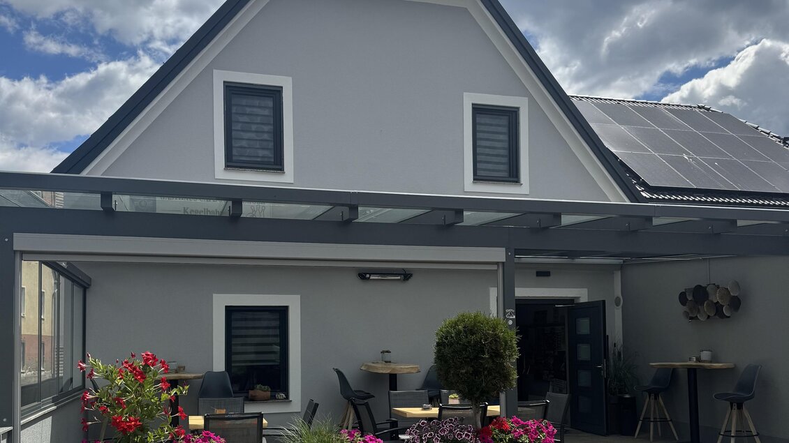 Ein modernes Haus mit einem grauen Anstrich und einem Pultdach. Vor dem Haus befinden sich blühende Pflanzen und eine überdachte Terrasse. | © Andrea Steinberger