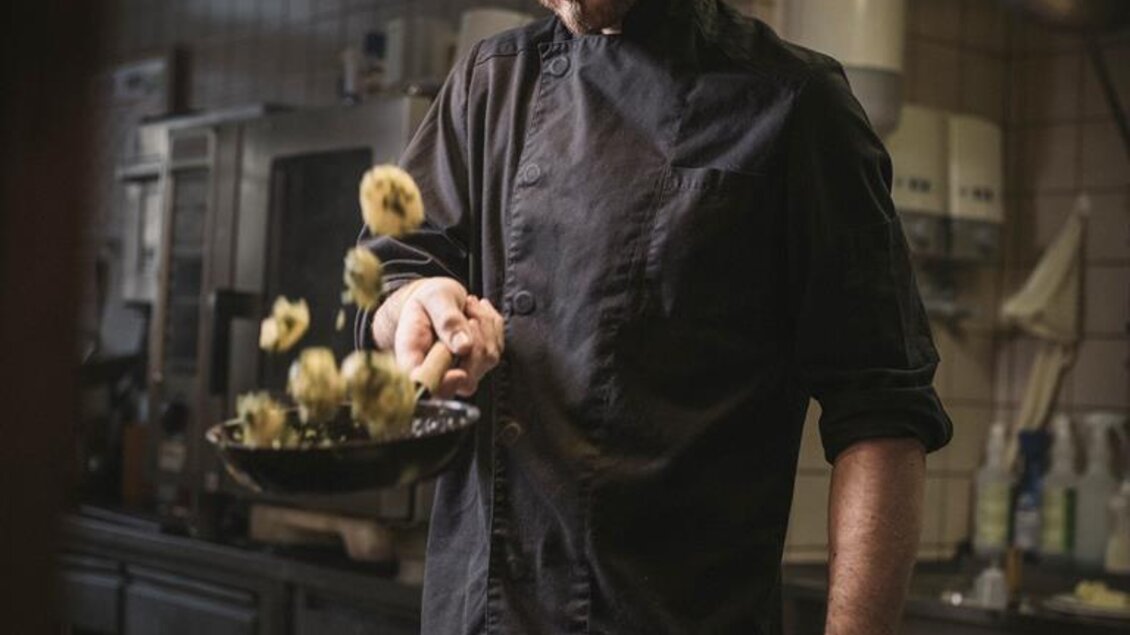 Ein Koch steht in einer Restaurantküche und brät Gemüse in einer Pfanne. Im Vordergrund liegt ein Teller mit Karottenstücken. | © Stefan Leitner