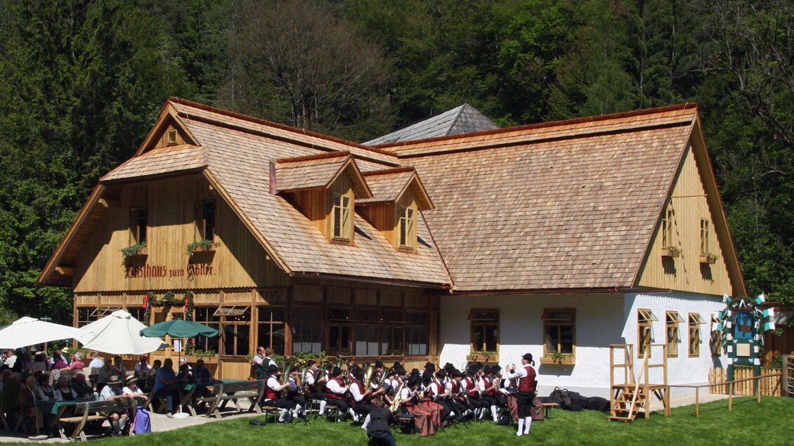 Ein traditionelles Haus inmitten der Natur, umgeben von Bäumen. Vor dem Haus sitzt eine Gruppe von Menschen in Trachten und genießt die Atmosphäre. | © OeFM Stuebing