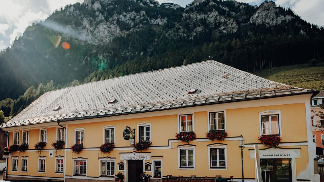 Ein gelbes Gebäude mit Blumenkästen, umgeben von Bergen. Es scheint ein Hotel oder eine Gaststätte zu sein. | © GenussReich