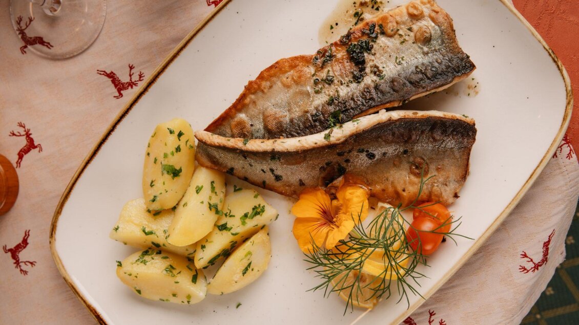 Ein Teller mit gebratenem Fisch, serviert mit Kartoffeln und frischen Kräutern. Daneben liegen essbare Blumen und Cherry-Tomaten. | © Gasthof Käferhube