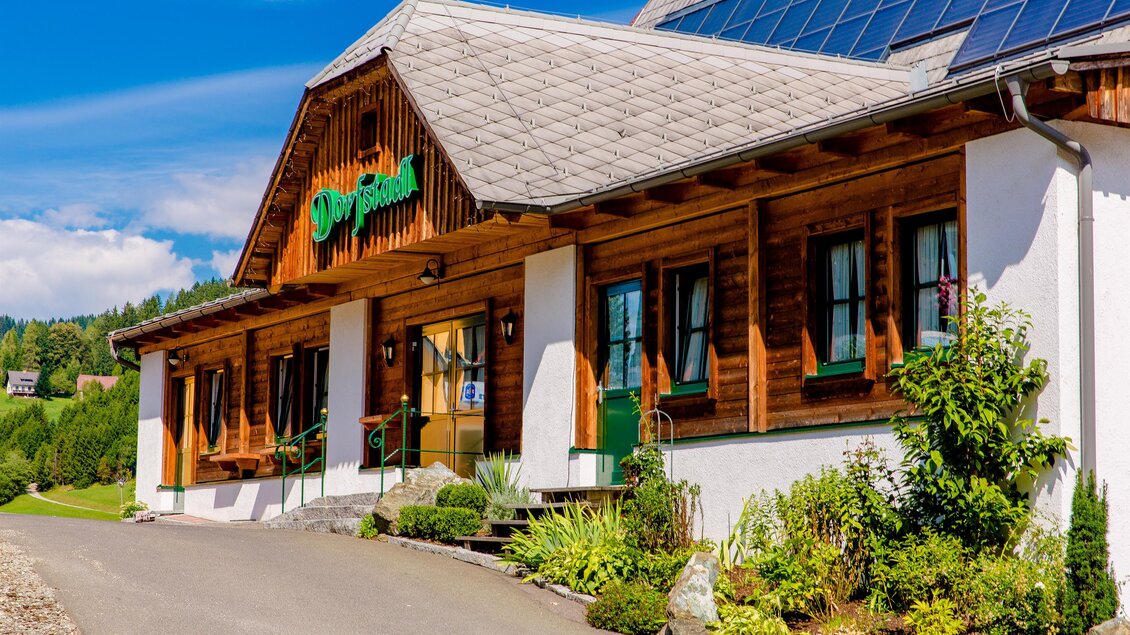 Ein traditionelles Holzhaus mit einem begrünten Eingang. Der Himmel ist klar und die Umgebung ist grün und einladend. | © Gasthof Landhotel Hubmann
