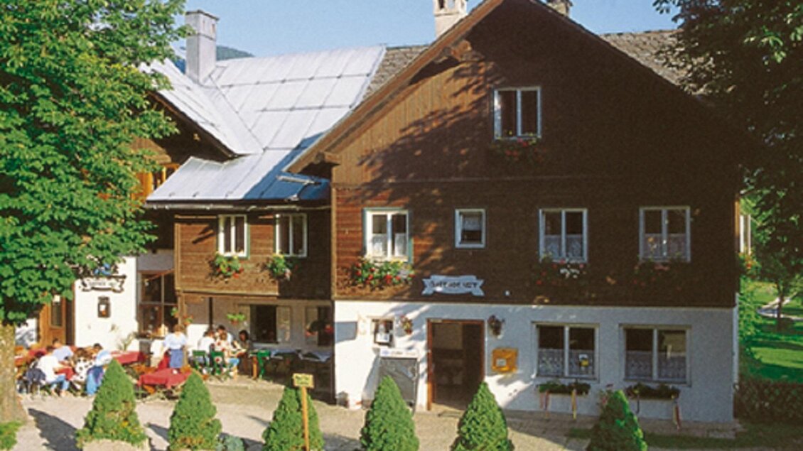 Ein traditionelles Holzhaus mit grünen Bäumen und blühenden Blumen. Gäste sitzen im Freien und genießen die Atmosphäre. | © Gasthof Veit