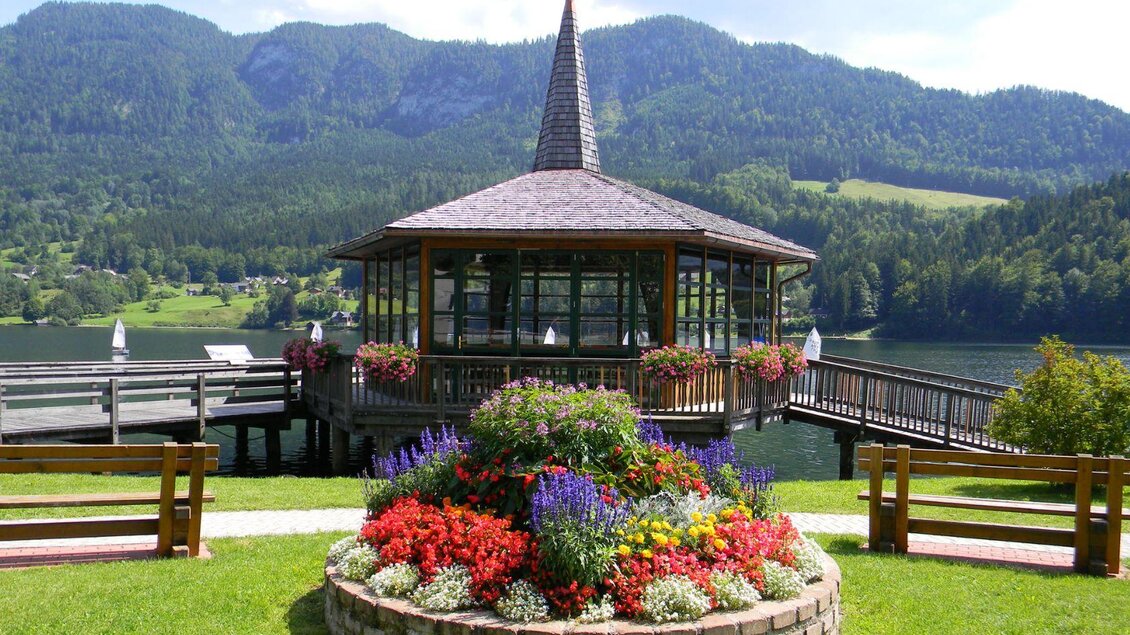 <p>Der Musikpavillon von Grundlsee direkt am Wasser, umgeben von farbenprächtigen Blumenbeeten. Im Hintergrund erheben sich die sattgrünen Berge unter einem strahlend klaren, blauen Himmel.</p> | © Gemeinde Grundlsee