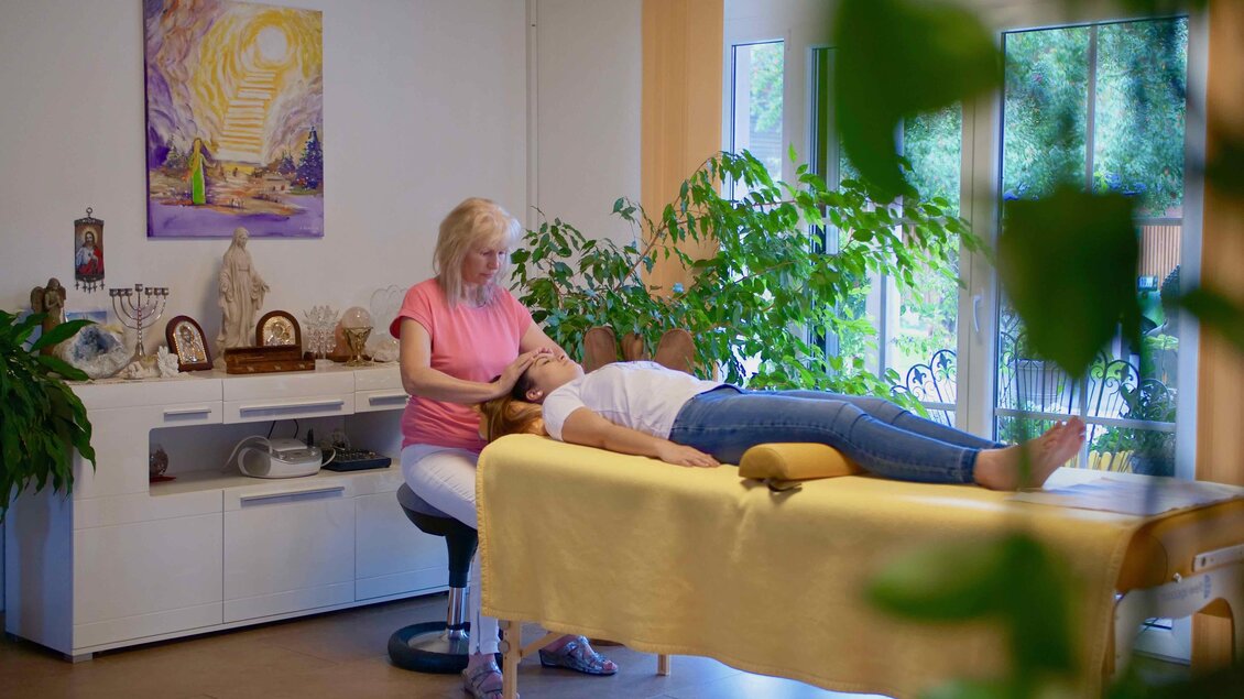 Eine Frau liegt entspannt auf einer Liege, während eine andere Frau ihr eine Behandlung gibt. Der Raum ist hell und freundlich, umgeben von Pflanzen und dekorativen Elementen. | © Fam. Offenbacher