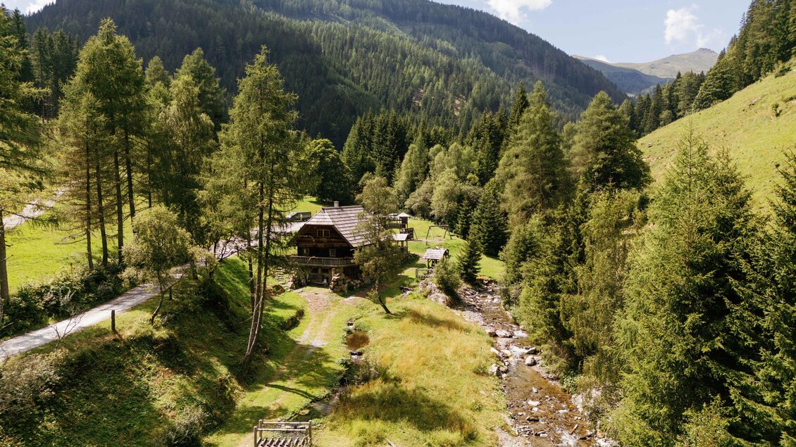 Eine idyllische Landschaft mit Bergen und dichten Wäldern. Ein kleines Haus liegt malerisch am Rand eines Baches. | © Karl Steinegger