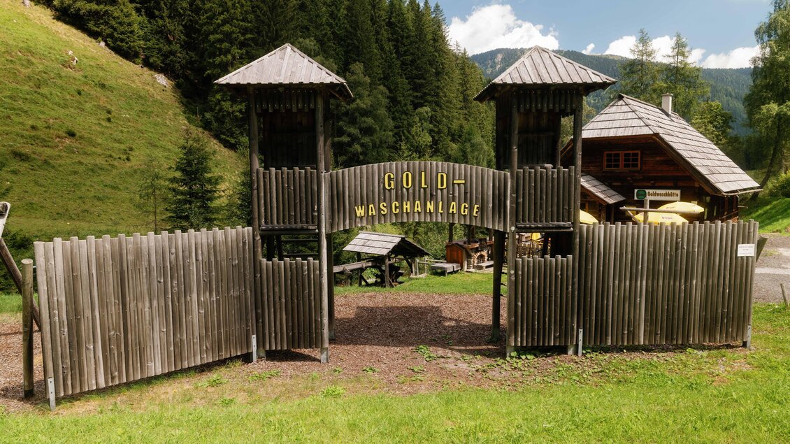Ein Eingang zu einer Goldwaschanlage, umgeben von Bäumen und Hügeln. Im Hintergrund steht ein traditionelles Holzhaus. | © Karl Steinegger