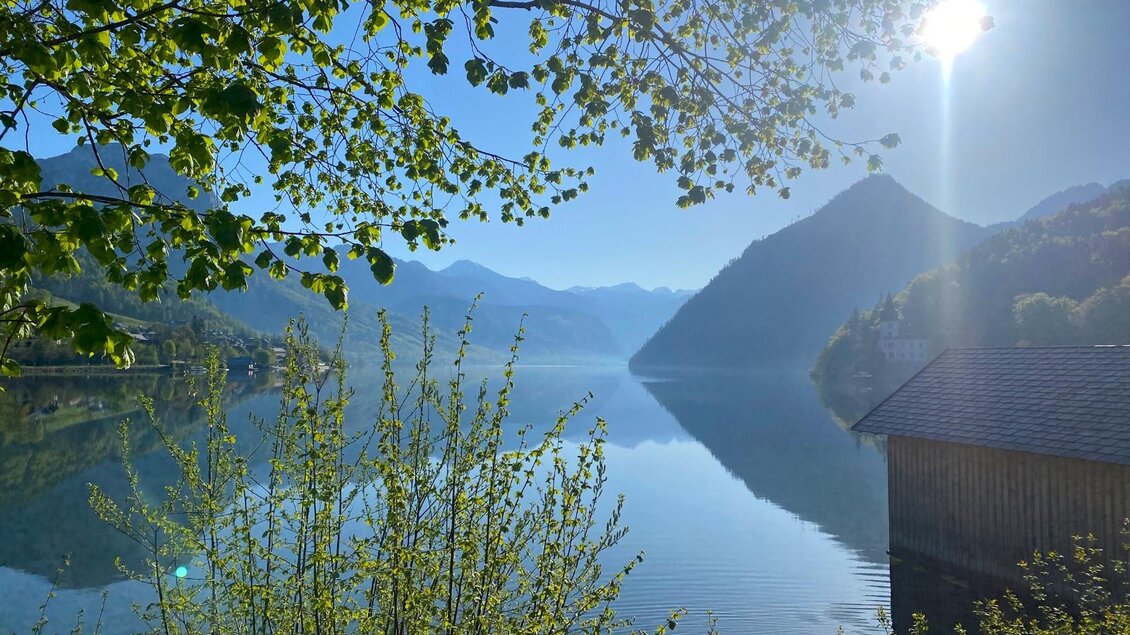Ein ruhiger See umgeben von Bergen und Bäumen. Die Sonne scheint hell am klaren blauen Himmel. | © Bettina Scheck