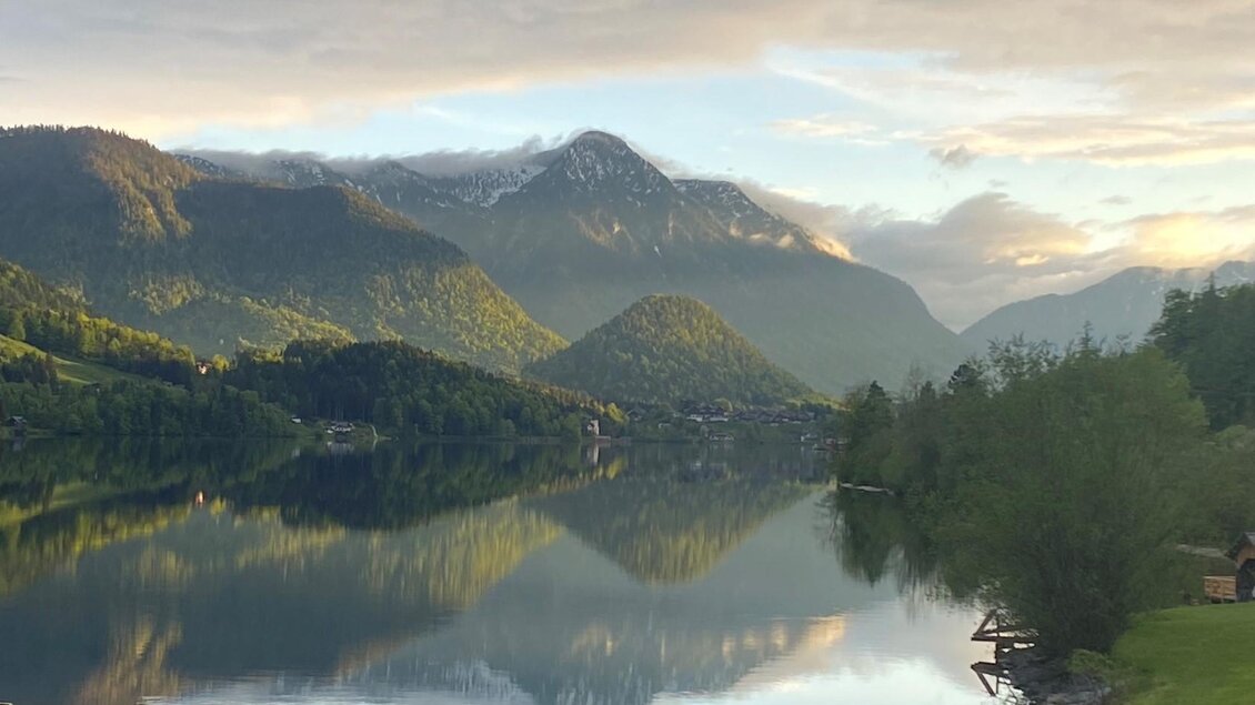 Ein ruhiger See umgeben von Bergen und üppigem Grün. Die Wolken spiegeln sich sanft im Wasser und schaffen eine friedliche Atmosphäre. | © Bettina Scheck