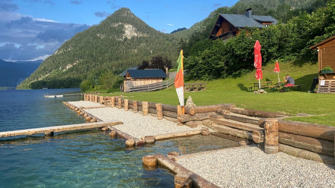 Ein malerischer See mit einem Steg aus Holz und Grasflächen. Im Hintergrund sind Berge und einige gemütliche Hütten zu sehen. | © Bettina Scheck