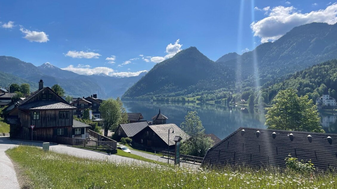 Eine malerische Landschaft mit Holzhäusern am Ufer eines Sees. Im Hintergrund erheben sich grüne Berge unter einem blauen Himmel. | © TVB Ausseerland/Bettina Scheck