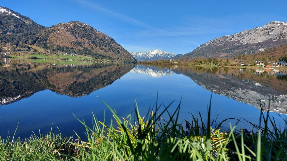Der sommerliche Grundlsee aus Richtung Gößl fotografiert, umgeben von Bergen des Ausseerland Salzkammergutes. Der Himmel ist klar und blau, und das Wasser spiegelt die wunderschöne Landschaft Grundlsees wider. | © Steinegger