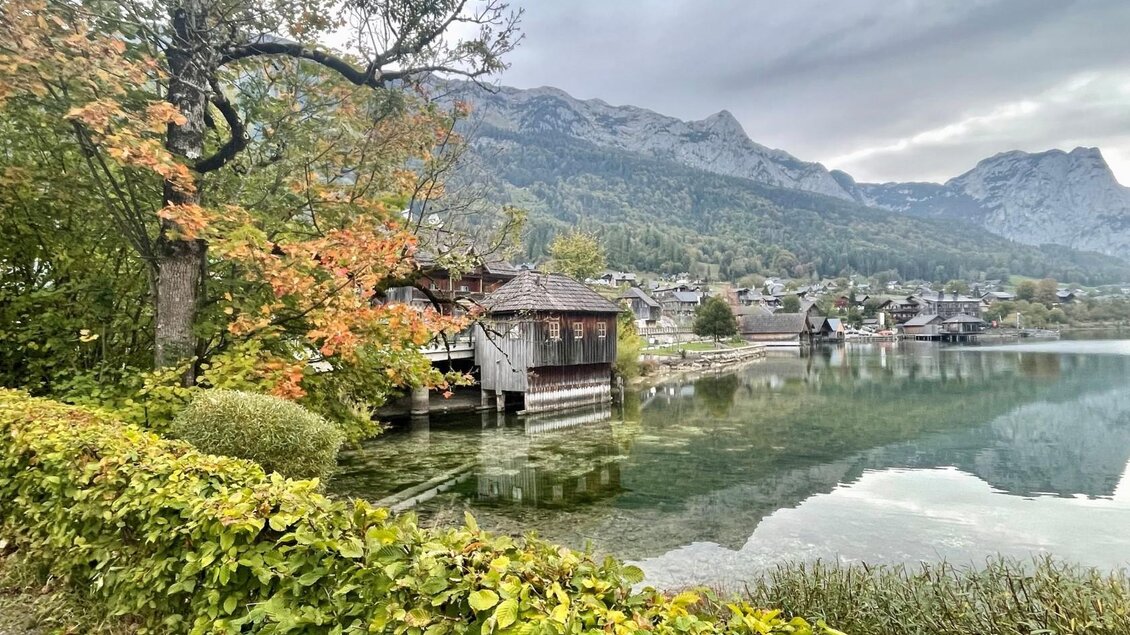 Ein malerischer See umgeben von Bergen und buntem Herbstlaub. Kleine Holzhäuser spiegeln sich im klaren Wasser. | © Bettina Scheck