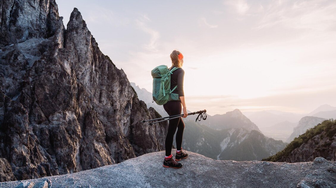 Ein Wanderer steht auf einem Felsen und schaut auf die bergige Landschaft. Die Sonnenstrahlen scheinen am Horizont und verleihen der Szene eine friedliche Atmosphäre. | © TV Gesäuse,