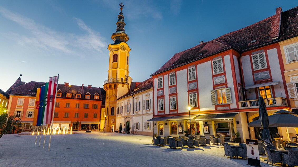 Historischer Hauptplatz von Bad Radkersburg in der Abenddämmerung mit beleuchtetem Kirchturm, bunten Häuserfassaden, Fahnen und Gastgärten. | © Zehnerhaus Bad Radkersburg