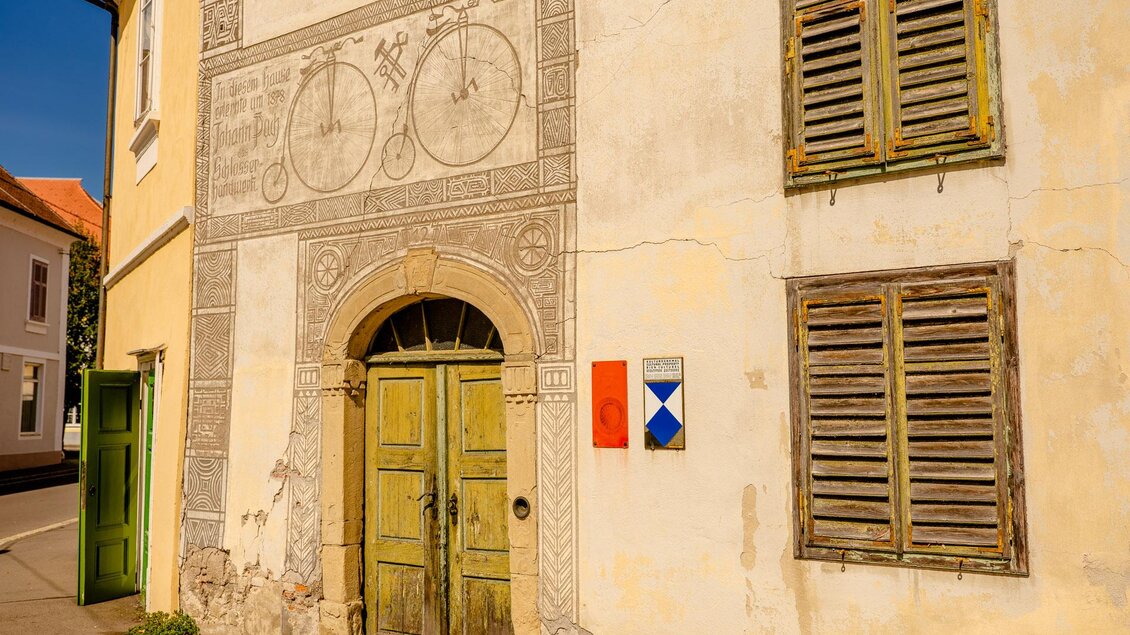 Historische Hausfassade in Bad Radkersburg mit altem Rundbogenportal, Holzläden und einer Wandzeichnung, die an Johann Puch und historische Fahrräder erinnert. | © Zehnerhaus Bad Radkersburg