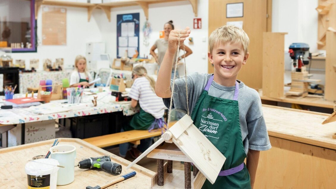 Ein Junge hält stolz ein selbstgebautes Vogelhaus in der Hand. Im Hintergrund arbeiten weitere Kinder an ihren Projekten. | © www.zloam.at