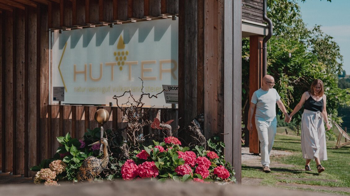 Ein Paar spaziert Hand in Hand am Eingangsbereich des Weinguts Hutter in Feldbach vorbei. Das Holzgebäude mit Weingut-Schild und blühenden Hortensien liegt im Thermen- & Vulkanland in der Steiermark. | © Thermen- & Vulkanland