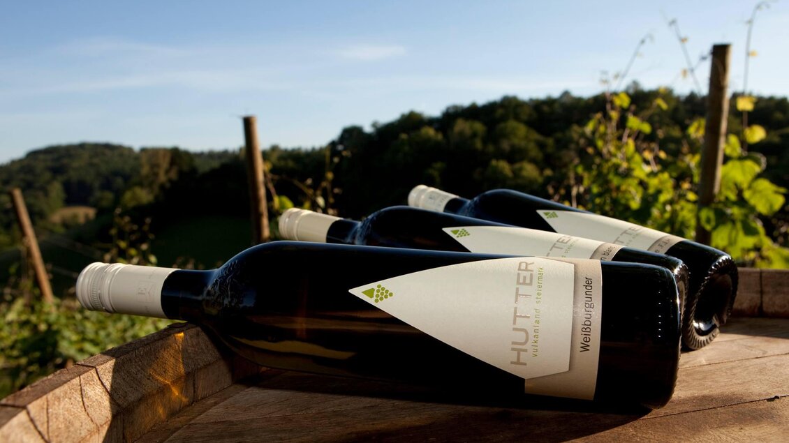 Drei Weinflaschen stehen auf einem rustikalen Tisch im Weinberg. Im Hintergrund erstrecken sich sanfte Hügel unter einem klaren Himmel. | © Weingut Hutter