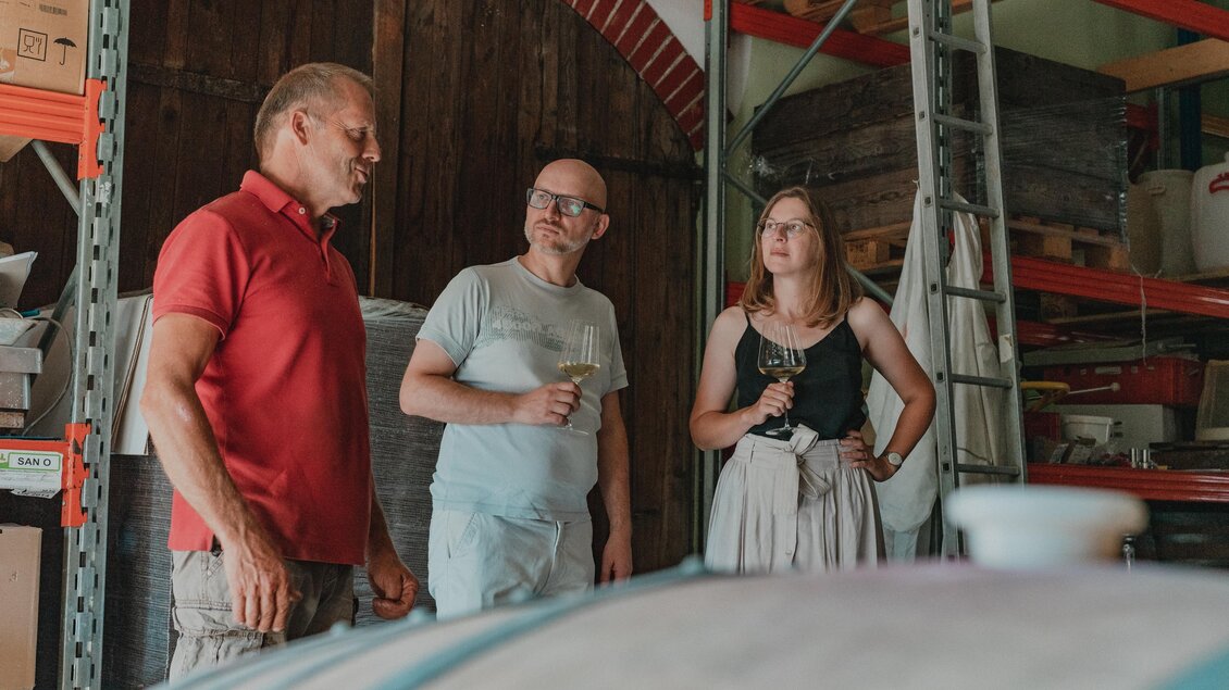 Drei Personen bei einer Weinverkostung im Weingut Hutter in Feldbach. Zwischen Holzfässern und Lagerregalen erhalten Gäste Einblicke in die Weinkultur im Thermen- & Vulkanland. | © Thermen- & Vulkanland
