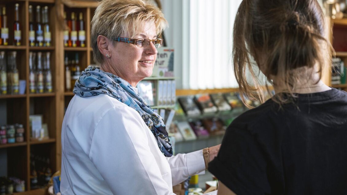 Eine Frau mit blonden Haaren und Brille spricht mit einer anderen Person. Im Hintergrund sind Regale mit verschiedenen Produkten zu sehen. | © Thomas Sattler
