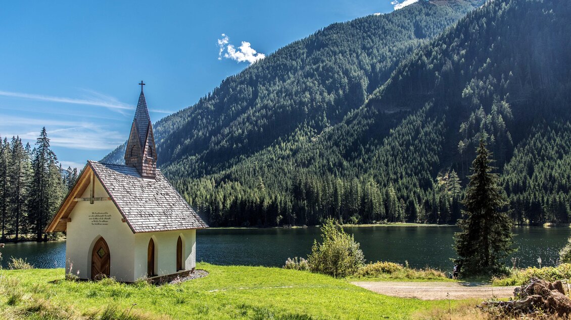 Eine kleine Kirche steht am Ufer eines klaren Sees, umgeben von hohen Bergen und dichten Wäldern. Der Himmel ist blau mit wenigen Wolken. | © Anita Fössl
