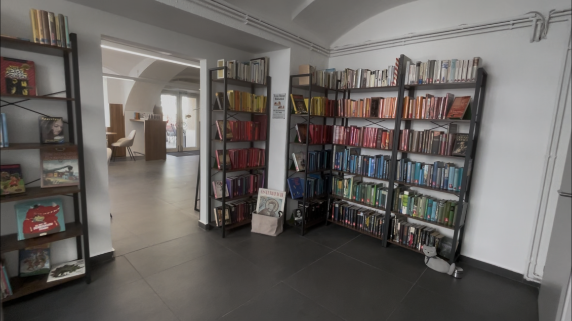 Ein moderner Raum mit Bücherregalen, die farblich sortierte Bücher zeigen. Im Hintergrund ist ein weiterer Bereich mit Sitzgelegenheiten sichtbar. | © Michaela Grasser