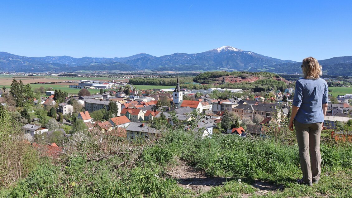 Eine Frau steht auf einem Hügel und blickt auf eine Landschaft mit einem kleinen Dorf im Tal. Im Hintergrund sind Berge sichtbar und der Himmel ist klar und blau. | © Weges