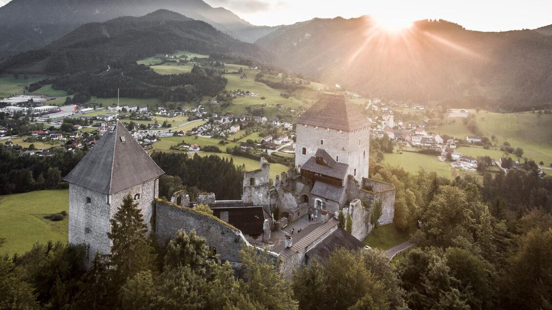 Eine alte Burg umgeben von grünen Hügeln und Wäldern. Die Sonne geht hinter den Bergen unter und taucht die Landschaft in warmes Licht.