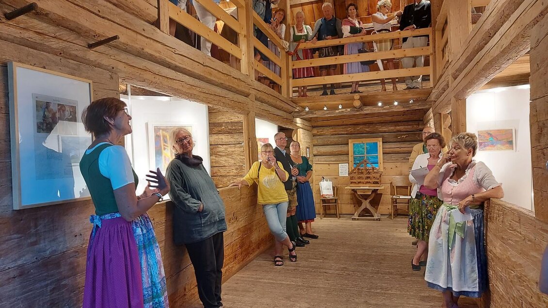 Eine Gruppe von Menschen hört einer Frau zu, die in einem Holzraum spricht. Der Raum ist mit Bildern dekoriert, und einige Personen stehen auf einer oberen Galerie. | © Kulturelle ARGE Grundlsee