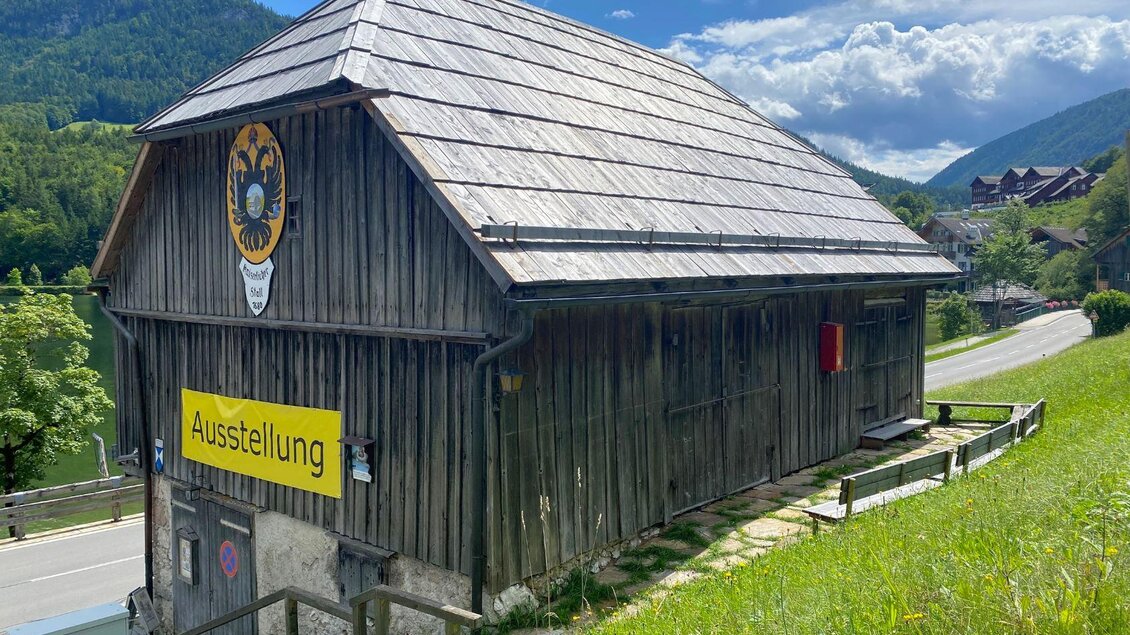 Ein traditionelles Holzgebäude mit einem großen Wappen und einem Schild, das „Ausstellung“ anzeigt. Im Hintergrund sind grüne Hügel und ein blauer Himmel zu sehen. | © TVB Ausseerland Salzkammergut/Bettina Scheck