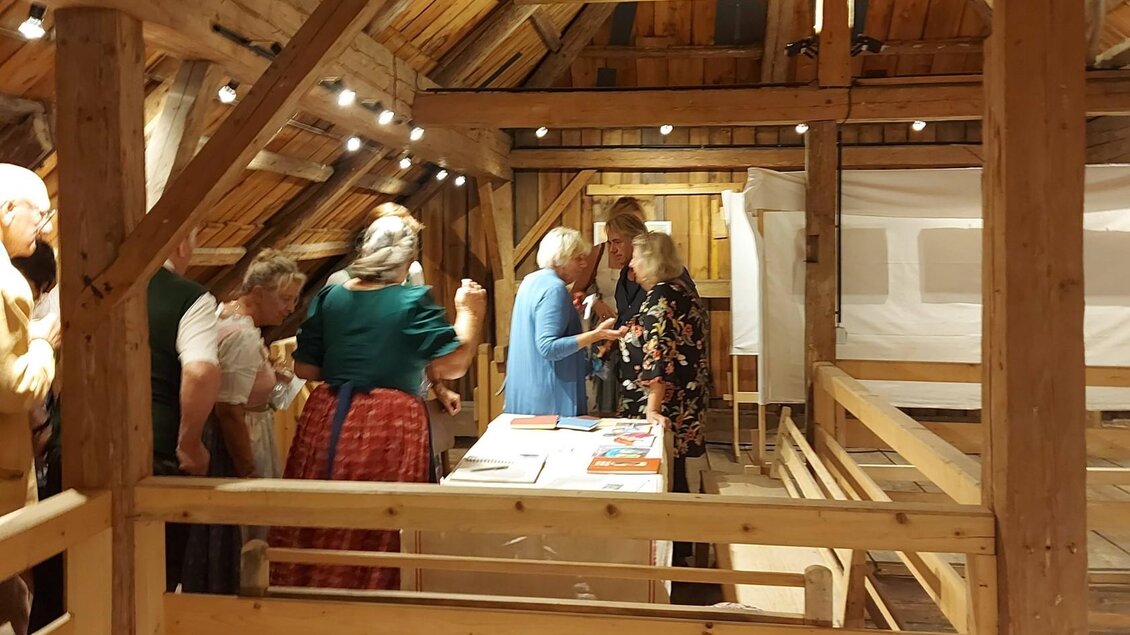 Eine Gruppe von Menschen steht in einem rustikalen Raum mit Holzdecke. Sie scheinen sich zu unterhalten und am Tisch mit Informationen aktiv zu sein. | © Kulturelle ARGE Grundlsee