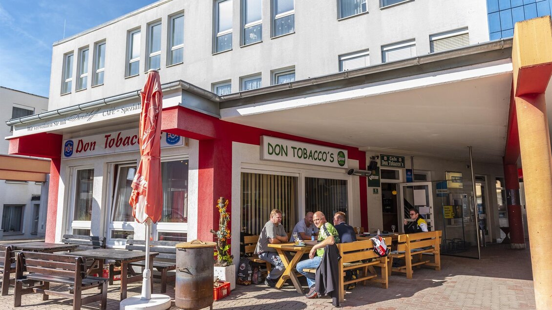 Ein Café mit Außenbereich, in dem Menschen an Tischen sitzen und sich unterhalten. Die Fassade des Cafés ist in Weiß und Rot gehalten, mit auffälligen Schirmen. | © Erlebnisregion Murtal