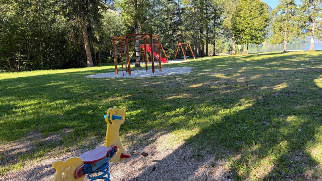 Der Spielplatz am Freizeitzentrum in Gößl am Grundlsee mit einer roten Rutsche und Schaukeln steht im Grünen. Im Hintergrund sind Bäume und der ruhige Grundlsee zu sehen. | © TVB Ausseerland Salzkammergut
